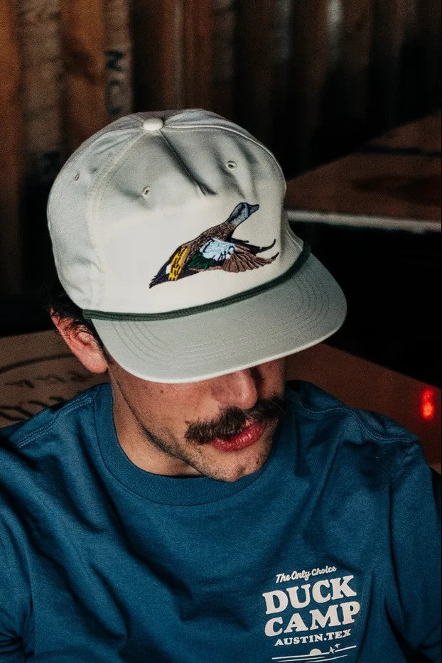 A man wearing a light gray baseball cap with an embroidered duck logo and a blue t-shirt with 'Duck Camp' printed on the front, set against a rustic wooden background.