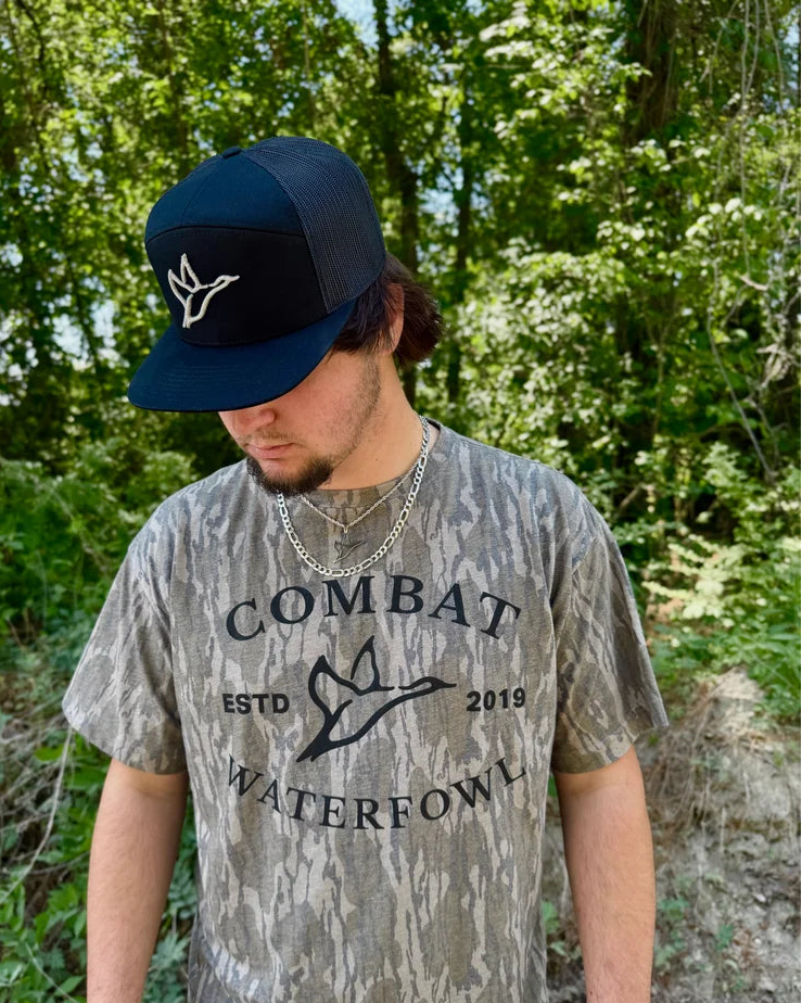 Person wearing a camouflage t-shirt with 'Combat Waterfowl' text and a navy cap in a forest setting.