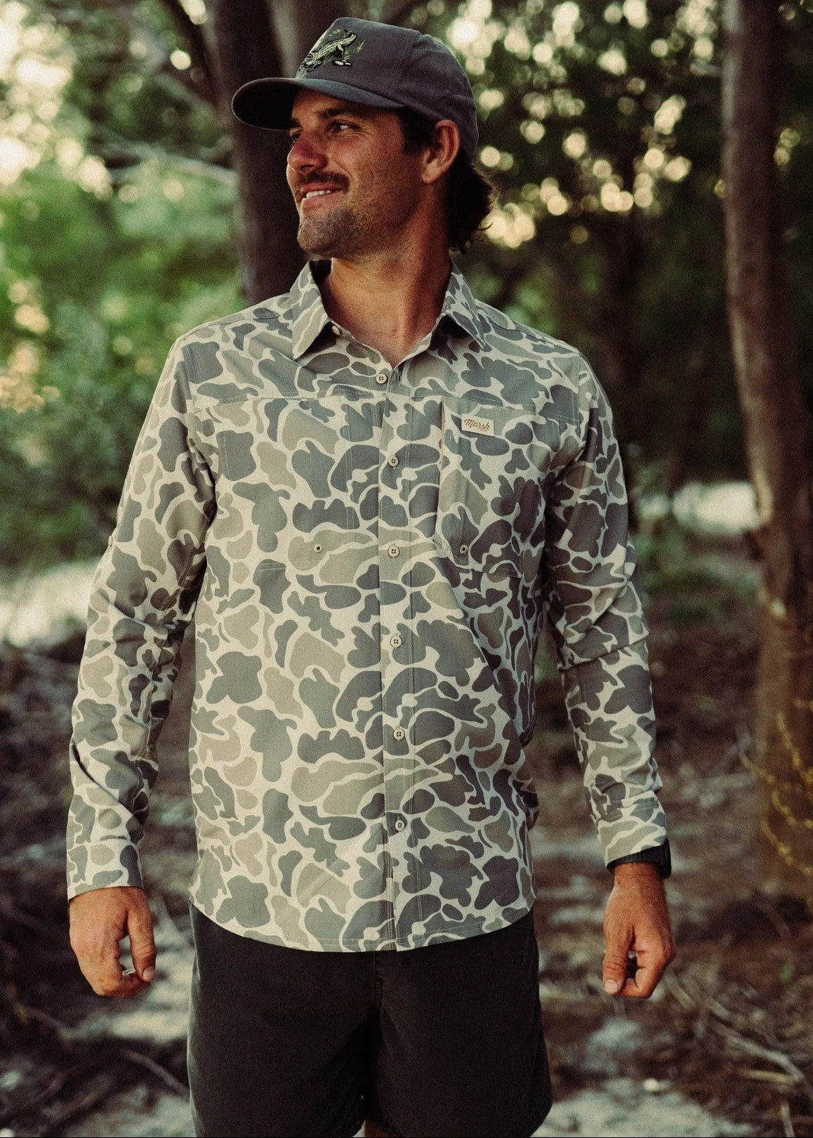 Man wearing a camouflage shirt and cap standing in a forest setting