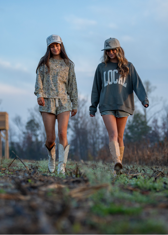 Two women walking outdoors in a field at dusk, wearing matching western-style outfits. One wears a beige camouflage long-sleeve top and shorts with white cowboy boots, and the other wears a dark gray sweatshirt with 'LOCAL' printed on the front, striped shorts, and beige boots.